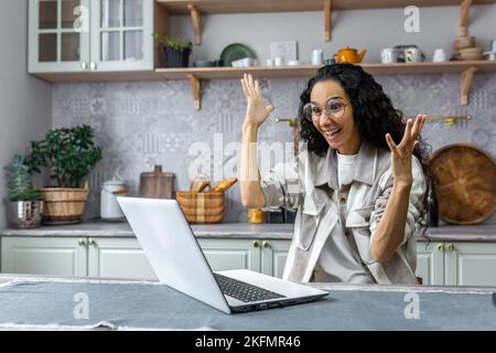Positive and creative Latin American woman sitting at home at the kitchen table and working, studying at the laptop. He talks to the camera, explains with his hands, smiles Stock Photo