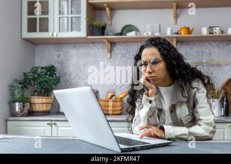 Boring and unmotivated online learning. Sad young beautiful female student sitting in the kitchen at home with a laptop, looking at the camera, holding her head with her hand Stock Photo