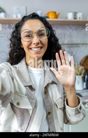 Young beautiful hispanic woman in glasses and beige shirt writes a blog, conducts an online stream. Talking to the camera at home in the kitchen. She smiles happily and waves her hand. Stock Photo