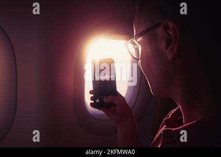 Portrait of man while taking pictures through airplane window. Passenger using phone during flight at beautiful sunset. Stock Photo