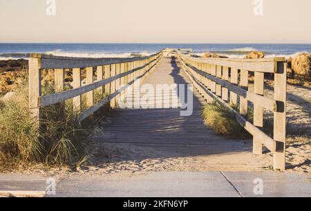 Wooden pier with grass on Atlantic ocean coast, Portugal. Wooden boardwalk to sea with rocks on shore. Empty walkway in dunes at dawn. Stock Photo