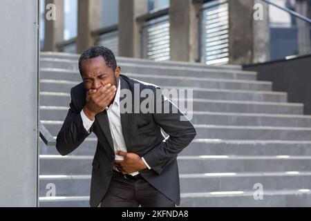 Nausea and pain in the stomach, a businessman from outside the office building is sick, a man is holding his hand on his stomach in a business suit, he got sick with poor-quality food. Stock Photo