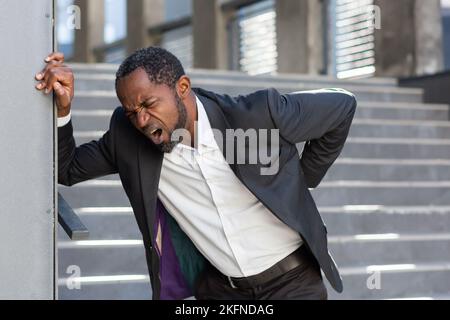 African american man overworked outside office building, boss in business suit has severe back pain, businessman near stairs holding hands on back muscles. Stock Photo