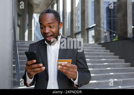 Happy african american businessman outside office building using phone and bank credit card for online shopping and money transfer, mature boss in business suit smiling. Stock Photo