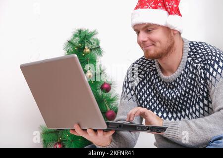 Man with laptop working, studying remotely new year 2023, Christmas. Man in Santa hat on background Christmas tree, garlands, gifts. Remote work. Surfing Internet. Man congratulates on holiday online. Stock Photo