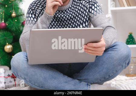 Man with laptop working, studying remotely new year 2023, Christmas. Man in Santa hat on background Christmas tree, garlands, gifts. Remote work. Surfing Internet. Man congratulates on holiday online. Stock Photo