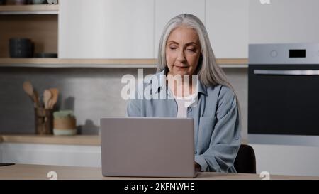 Elderly businesswoman female senior freelancer work on laptop from home typing on laptop keyboard chatting online use computer software app browsing Stock Photo