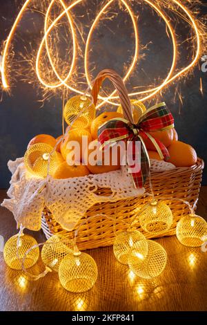 Tangerines and garlands lie in a basket on a blue background. In the photo, the light lines of sparklers. There is a red bow on the basket. Stock Photo