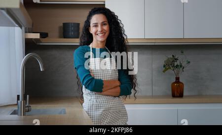 Smiling young woman maid housewife mistress baker housekeeper with long curly hair wearing checkered apron standing with folded arms confident chef Stock Photo