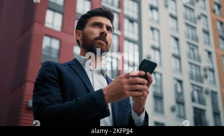 Caucasian bearded man entrepreneur businessman hold cellphone texting messages stand in city with modern office buildings use mobile business Stock Photo