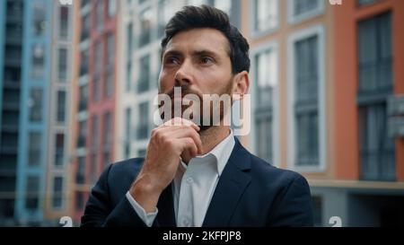 Caucasian smart puzzled thoughtful man hold hand to chin dreaming about career growth successful employer in stylish black suit stand outside modern Stock Photo