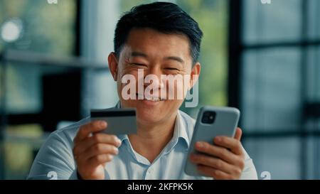 Happy Asian man client buyer businessman use banking mobile phone app at office hold plastic credit card enter secure code buy goods in internet store Stock Photo
