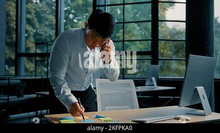 Busy asian businessman korean man working stand at table in modern office talk on phone give professional consultation by mobile call write tasks on Stock Photo