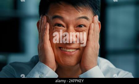 Positive emotional asian korean man businessman 40s male employee hold face by hands feel happiness triumph good shock opens mouth with delight wow Stock Photo