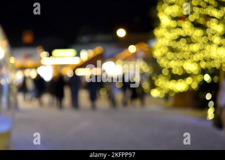 Blurred background. Christmas tree, decorated glowing lights, building, blurry silhouettes of people walking city square at winter night. Beautiful New Year and Christmas holiday blurred background Stock Photo