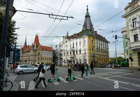 Cluj-Napoca, Romania - September 17, 2022: Young people on electric scooters cross the street near Szeki Palace, left, and Babos Palace, right, in Clu Stock Photo