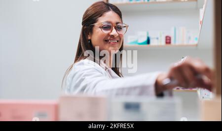 Pharmacy worker getting medication from a shelf. Indian female pharmacist dispensing a prescription in a chemist. Happy woman working in a drug store. Stock Photo