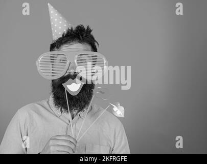 Guy in party hat celebrate, posing with photo props. Hipster in giant sunglasses celebrating. Man with beard on cheerful face holds smiling lips on Stock Photo