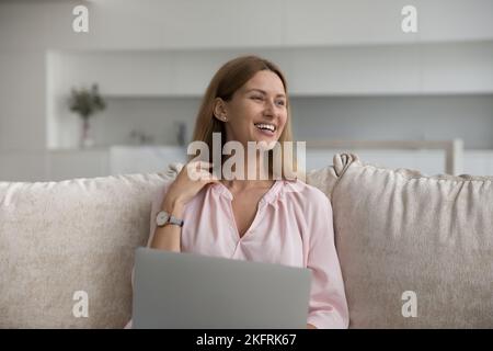Smiling young woman sitting on couch at home using laptop Stock Photo