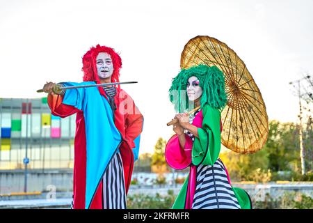 Full length of creative talented performers with face art in colorful costumes and wigs standing on stilts in park with Asian umbrella and sword during show, on sunny day Stock Photo