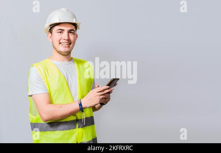 Smiling engineer using cell phone looking at camera. Friendly male builder engineer using phone and looking at camera. Young engineer in vest and helm Stock Photo