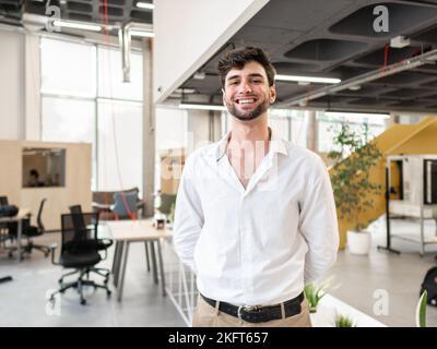 Smiling young bearded man in smart casual outfit standing near table in modern spacious workspace and looking at camera Stock Photo