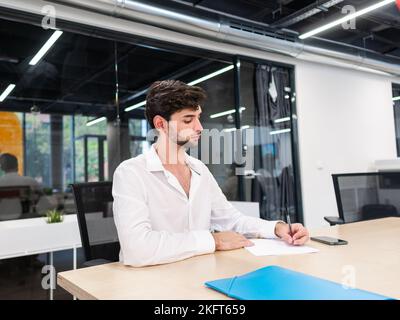 Serious bearded young man sitting at table with blue folder in modern office and taking notes during workday Stock Photo