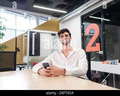 Happy young bearded man in white shirt sitting at table and texting message via cellphone in light modern workspace Stock Photo