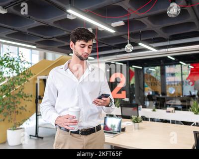 Young bearded man in smart casual outfit standing near table with takeaway coffee and browsing on smartphone while looking at screen in modern workspa Stock Photo
