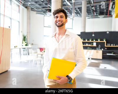 Smiling young bearded businessman in white shirt standing in light spacious office cafeteria and holding colorful folders while looking at camera Stock Photo