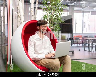 Happy young male entrepreneur in smart casual outfit sitting on modern chair and typing information via laptop in modern workspace with green plant Stock Photo
