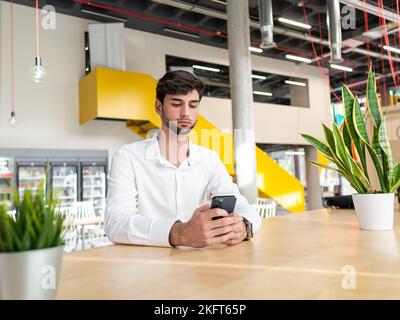 Concentrated young bearded man in white shirt sitting at table and texting message via cellphone in light modern workspace Stock Photo