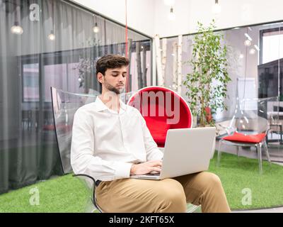 Concentrated young male entrepreneur in smart casual outfit sitting on transparent chair and typing information via laptop in modern workspace with gr Stock Photo