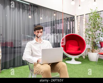 Happy young male entrepreneur in smart casual outfit sitting on transparent chair and typing information via laptop in modern workspace with green pla Stock Photo