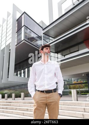 Low angle of pensive young bearded man in smart casual outfit standing near urban building with hands in pockets and looking away Stock Photo