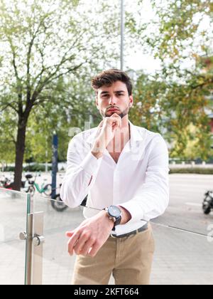 Pensive young bearded man in smart casual outfit and wristwatch standing near glass fence and touching chin while looking at camera against green tree Stock Photo