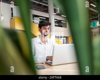 Focused young bearded businessman in white shirt sitting at table with smartphone and typing information on laptop in light spacious workspace Stock Photo