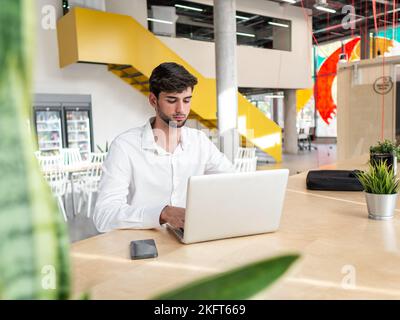 Focused young bearded businessman in white shirt sitting at table with smartphone and typing information on laptop in light spacious workspace Stock Photo