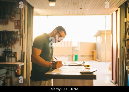 Concentrated male artisan writing on paper sheet while ordering parts at table in workshop Stock Photo
