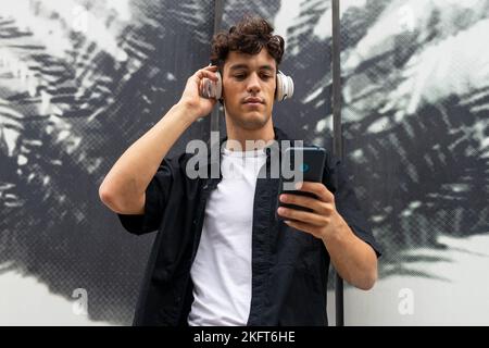 Young man in casual clothes listening to music from your smartphone while standing near wall with palm leaves ornament on city street Stock Photo