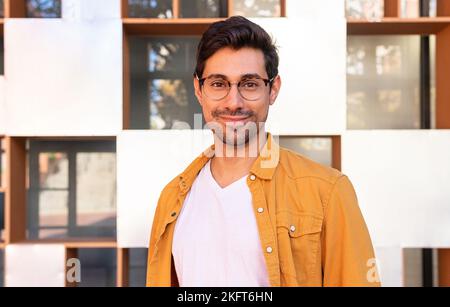 Positive young Hispanic bearded man in casual clothes and eyeglasses standing near wall of urban building and looking at camera Stock Photo