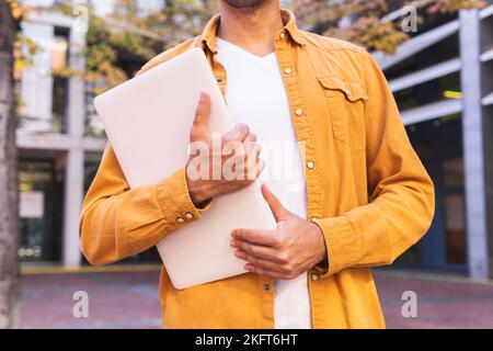 Cropped unrecognizable Hispanic bearded man in casual outfit and eyeglasses standing looking away near urban building with laptop Stock Photo