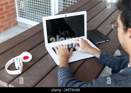 Cropped unrecognizable concentrated male student in eyeglasses and casual clothes sitting at wooden table near headset and typing information on lapto Stock Photo