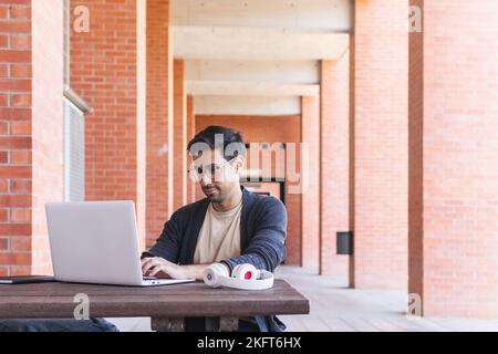 Concentrated bearded male student in eyeglasses and casual clothes sitting at wooden table near headset and typing information on laptop in modern wor Stock Photo