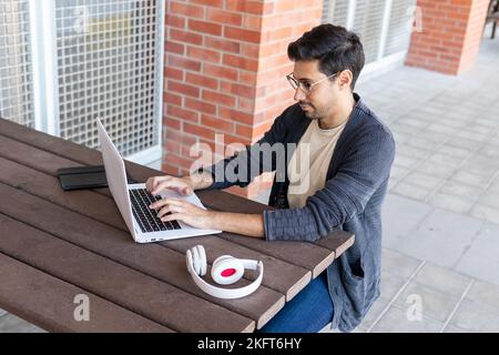 From above concentrated bearded male student in eyeglasses and casual clothes sitting at wooden table near headset and typing information on laptop in Stock Photo