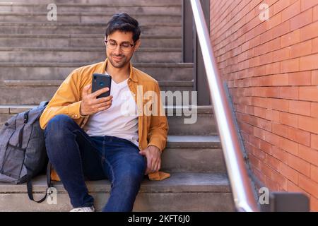 Focused young bearded Hispanic man in jeans and eyeglasses sitting on stone staircase with backpack and surfing internet via cellphone during daytime Stock Photo