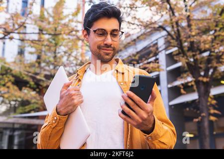 Positive young Hispanic bearded man in casual outfit and eyeglasses standing near urban building with laptop and browsing internet via cellphone Stock Photo