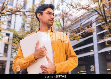 From below young Hispanic bearded man in casual outfit and eyeglasses standing looking away near urban building with laptop Stock Photo