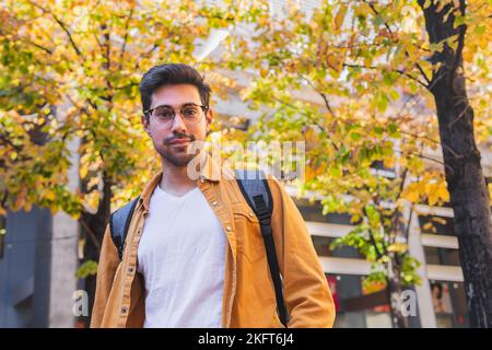 From below bearded male in casual outfit and eyeglasses with backpack standing near tall autumn trees looking at camera Stock Photo