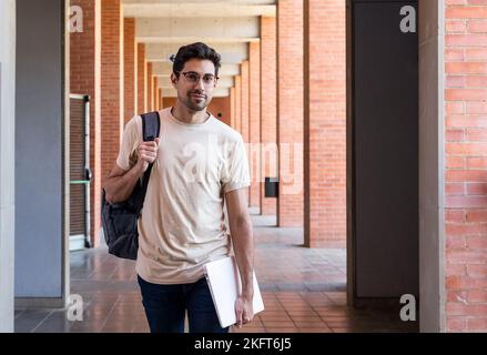 Positive young bearded ethnic man in casual outfit and eyeglasses holding laptop and backpack while walking along corridor with brick columns while lo Stock Photo